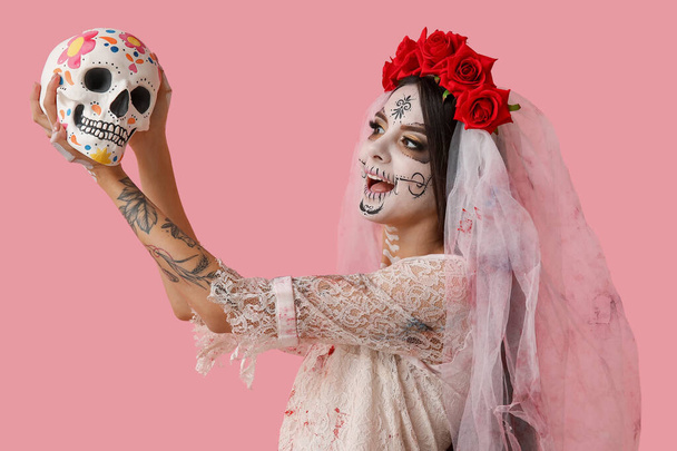 Cadılar Bayramı partisi için ölü gelin gibi giyinmiş genç bir kadın. Pembe arka planda insan kafatası resmi. - Fotoğraf, Görsel