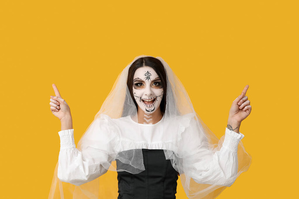 Cadılar Bayramı partisi için ölü gelin gibi giyinmiş güzel bir kadın sarı arka plandaki bir şeyi işaret ediyor. - Fotoğraf, Görsel