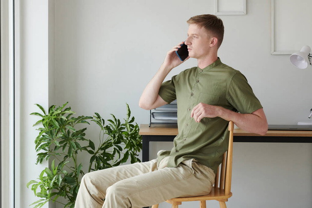 Νεαρός κάθεται στην καρέκλα μπροστά από το παράθυρο και μιλάει στο τηλέφωνο με συνάδελφο - Φωτογραφία, εικόνα
