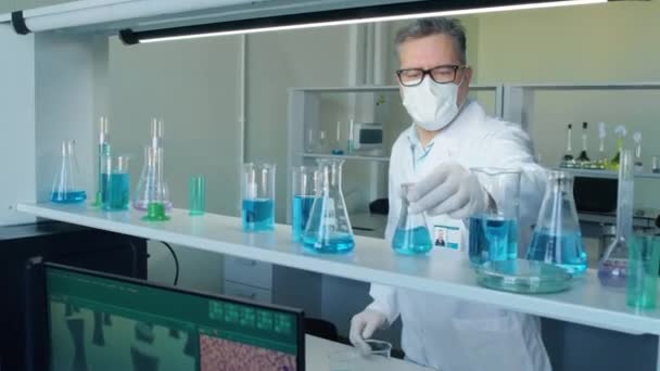 Medium shot of male senior scientist in mask taking flask with blue liquid from shelf and studying it - Footage, Video