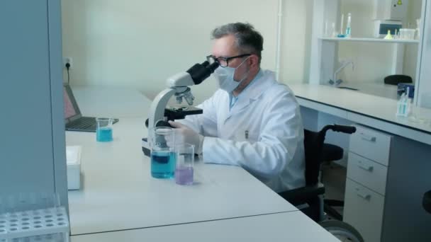 Középkorú férfi fehér tudós portréja, aki tolószékben ül az íróasztalnál, és a szondát nézi mikroszkóppal a laboratóriumban nappal - Felvétel, videó