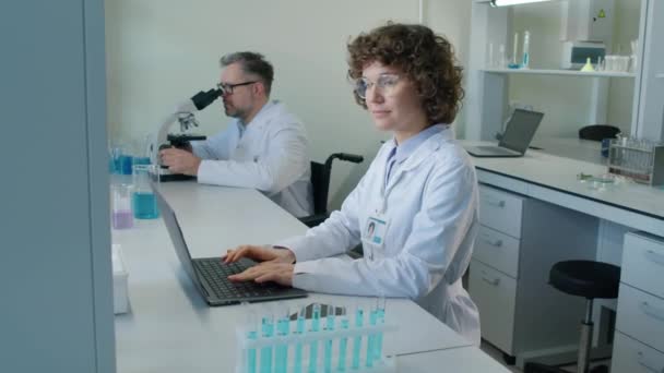 Retrato lento de la alegre mujer caucásica en bata médica blanca sentada frente a la computadora portátil en el laboratorio durante el día - Imágenes, Vídeo