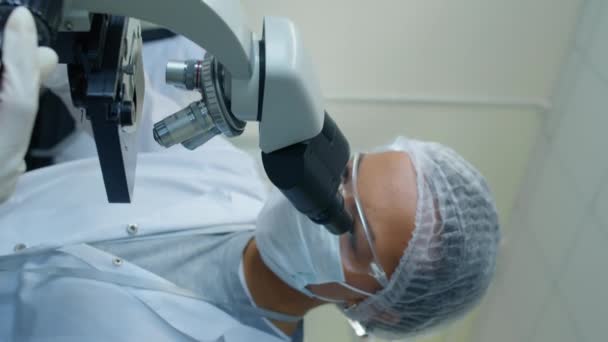 Вертикальный средний план женщины-ученого, носящей маску, очки и сетку для волос, регулирующей и смотрящей через микроскоп в лаборатории днем - Кадры, видео
