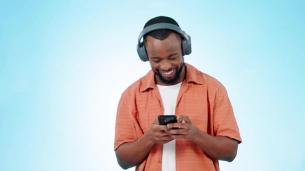 Black man, smartphone and headphones with dancing to music, listening to radio in studio on blue background. Dancer, fun and energy with rave or techno, audio streaming and happiness with mobile app. - Footage, Video