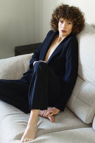 Cuerpo completo alto ángulo de hembra adulta con pelo rizado oscuro sentado en el sofá con chaqueta negra y pantalones mirando a la cámara - Foto, Imagen