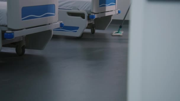 Sluiten van volwassen verpleegkundige dweilen vloer in de buurt van bedden in helder ziekenhuis afdeling. Gezondheidswerkers zorgen voor reinheid in het medisch centrum. Cleaner bereidt ziekenhuisruimte voor nieuwe patiënten. Medisch personeel aan het werk. - Video