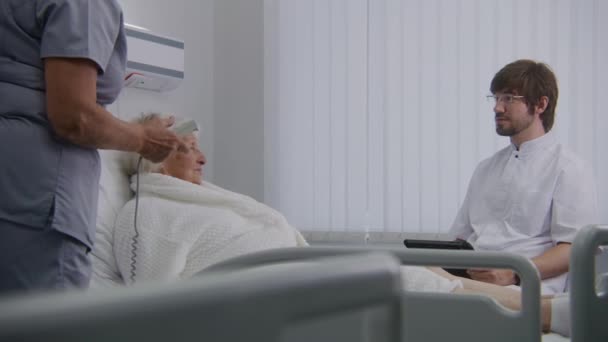 Doctor with tablet computer does checkup, consults female elderly patient resting in hospital ward. Nurse adjusts pillow and bed for senior woman recovering after successful surgery. Modern clinic. - Footage, Video