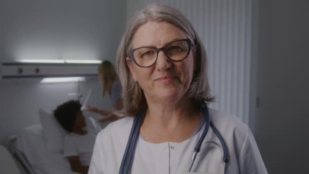 Felnőtt női orvos szemüvegben és sztetoszkóppal a nyakán áll a kórház közepén. A nővér ellenőrzi az afro-amerikai beteg állapotát a háttérben. A klinikán dolgozó egészségügyi személyzet. - Felvétel, videó