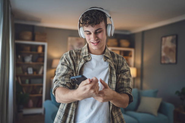 Πορτρέτο του έφηβος αγόρι σταθεί στο σπίτι χρήση ακουστικών και smartphone για να παίξετε μουσική ή να παρακολουθήσετε βίντεο podcast στο σπίτι - Φωτογραφία, εικόνα