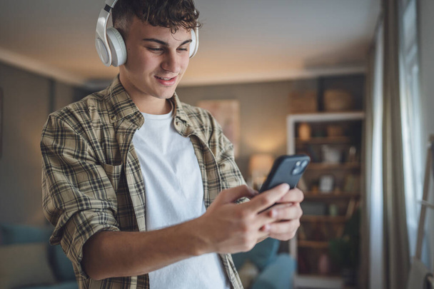 Portré tizenéves fiú állni otthon használja fejhallgató ans okostelefon zenélni, vagy nézni video podcast otthon - Fotó, kép