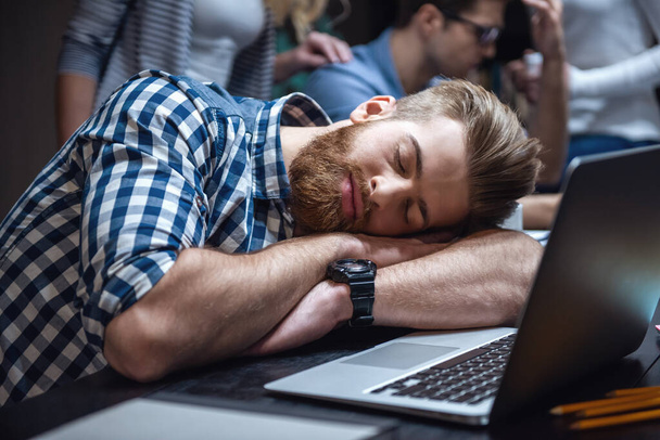 Knappe bebaarde man is slapen in de buurt van de laptop tijdens het werken hard in de bibliotheek - Foto, afbeelding