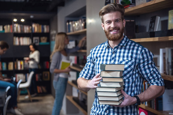 Los jóvenes inteligentes están trabajando duro en la biblioteca moderna. El tipo en primer plano sostiene un montón de libros, mira a la cámara y sonríe
 - Foto, imagen