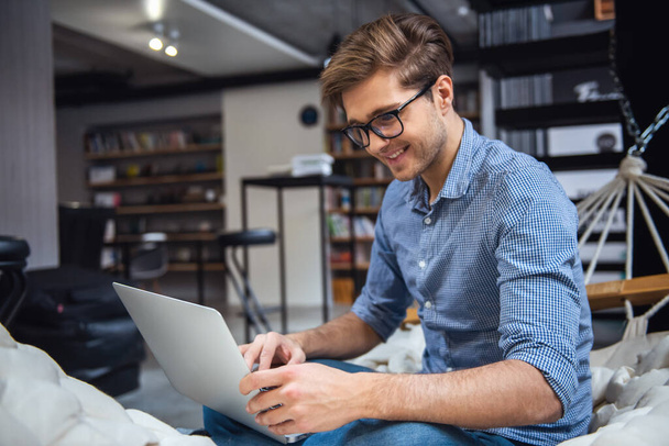 attraktiver junger Mann mit Brille benutzt einen Laptop und lächelt beim Lernen in der modernen Bibliothek - Foto, Bild