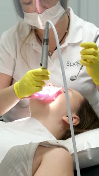 fogászat serdülőkor egy orvos sárga kesztyűben kitölti egy fiatal lány fogát egy nő közelkép videó a fogászat a fogorvosnál szárítja a fogat könnyű új technológiákkal. modern klinika - Felvétel, videó