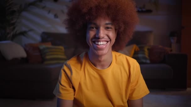 Widok z przodu wesoły młody afro człowiek podejmowania rozmowy wideo siedzi w salonie. Uśmiechnięty mężczyzna rozmawiający wieczorem przez internet w domu. Związki przyjaźni, aplikacje randkowe i sieci społecznościowe z - Materiał filmowy, wideo