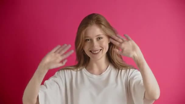 Eine junge europäische oder amerikanische Ingwerfrau in weißem T-Shirt posiert im Studio auf rosa Hintergrund. Ein fröhliches und fröhliches Mädchen mit einem schönen Lächeln wedelt mit den Armen und begrüßt das Publikum - Filmmaterial, Video