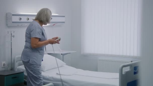 Mature nurse adjusts bed in hospital ward for patient using remote control. Female medical worker pushes wheelchair with old woman, puts her in bed. Medical staff in modern clinic or medical facility. - Footage, Video