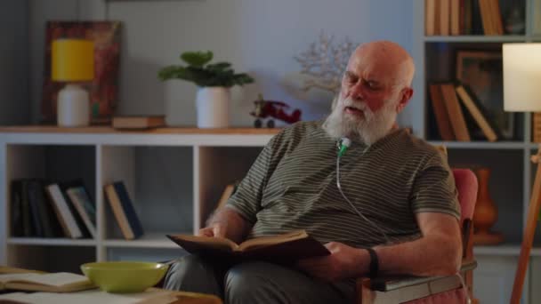 Vanha mies hengittää naamio pysyä tuolissa ja lukea kirjaa. Vanhus käyttää sumutinta vaikean hengityksen takia. Eri keuhkosairauksien lääketieteellisen kuraation aihe. Laadukas 4k kuvamateriaalia - Materiaali, video