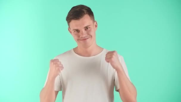 Portrait of a pleasantly excited young European guy in studio. A young man with a beautiful smile in a white T-shirt on a blue background shows a gesture of victory, satisfaction with himself in - Footage, Video