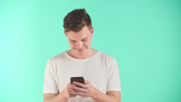 Young man is holding smartphone, using mobile apps, watching funny video in social media. Smiling man having fun chatting. Male has conversation with girlfriend on phone on blue background in studio - Footage, Video