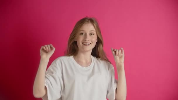Młoda Europejka lub amerykańska ruda w białej koszulce pozuje w studio na różowym tle. Ładna dziewczyna z pięknym uśmiechem tańcząca, nucąca, mówiąca, machająca rękami i dotykająca - Materiał filmowy, wideo