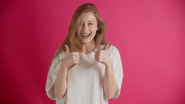 Молодая европейская или американская рыжая женщина в белой футболке позирует в студии на розовом фоне. Улыбающаяся счастливая и радостная девушка поднимает руки с поднятыми большими пальцами. Утверждение ведения бизнеса и - Кадры, видео