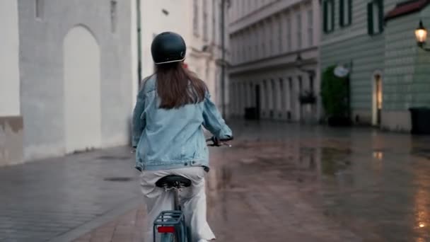 Vue arrière de la belle jeune femme à vélo le long de la rue pluvieuse au centre-ville. Concept de mode de vie actif - Séquence, vidéo