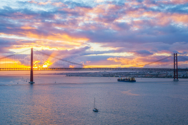 Widok 25 de Abril Bridge słynny zabytek turystyczny Lizbony nad rzeką Tagus z łodzi turystycznych i kontenerowiec ładunku na zachód słońca. Lizbona, Portugalia - Zdjęcie, obraz