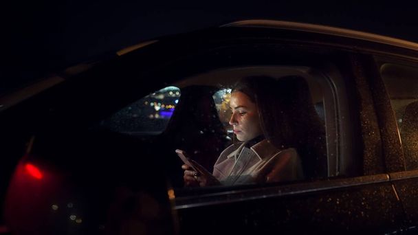 Zaostřená podnikatelka rolování mobilní telefon v řidiče sedí při dojíždění z kanceláře v luxusním autě v noci - Fotografie, Obrázek