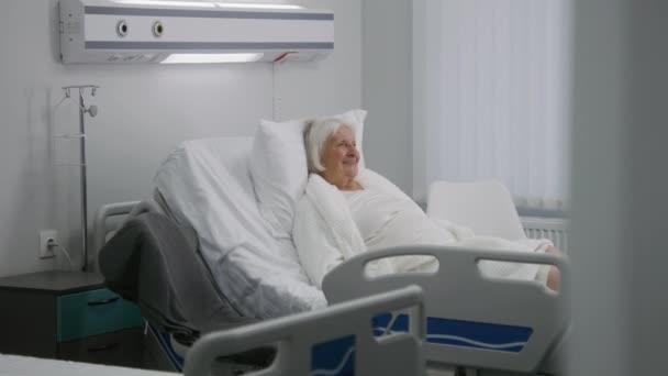 Elderly female patient rests in bed in bright hospital room. Loving family visits senior grandmother recovering after successful surgery, gives flowers. Modern medical facility or clinic. Slow motion. - Footage, Video