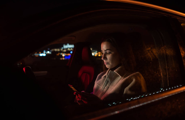 Ernstige zakenvrouw met behulp van smartphone in de bestuurder zit tijdens het rijden van het werk in luxe auto 's nachts. Stedelijke straat met werkende neon borden - Foto, afbeelding