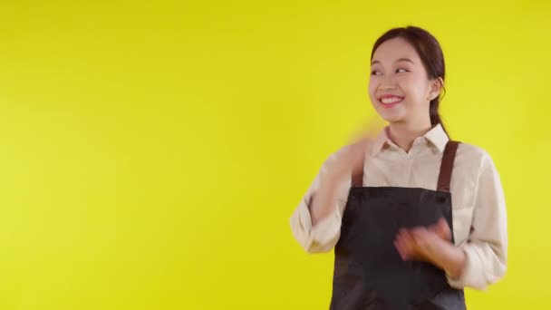 Retrato de joven barista asiática vestida de delantal presentando y pulgares arriba sobre fondo amarillo, camarera o emprendedora alegre y mostrando algo, pequeña empresa o startup, camarera de cafetería. - Metraje, vídeo