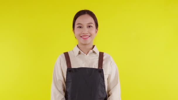 Önlük giymiş Asyalı genç bir baristanın portresi sarı arka planda gülümsüyor, garson ya da girişimci neşeli, küçük işletme ya da girişim sahibi, kendine güvenen bir garson ya da kafe garsonu. - Video, Çekim