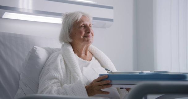 高齢の女性は美味しい食べ物を食べて,明るい病院区のベッドで眠っているカメラを見ています. 成功した手術後に回復する女性の高齢患者. 現代の医療施設やクリニック。 ポートレイト. - 写真・画像