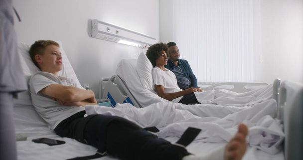 Hemşire bandajları yatağın üzerinde yatıyor çocuk. Afrikalı Amerikalı çocuk hastane koğuşunda dinleniyor ve babasıyla birlikte televizyon izliyor. Hasta başarılı bir ameliyattan sonra iyileşiyor. Modern, parlak tıp merkezi. - Fotoğraf, Görsel