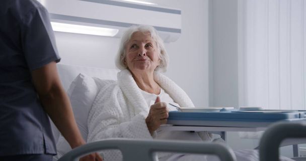 Infirmière ouvre une grande boîte à lunch avec repas à une patiente âgée. Femme âgée se reposant au lit dans une chambre d'hôpital lumineuse, se rétablissant après une chirurgie réussie, mangeant de la nourriture. Installation médicale ou clinique moderne. - Photo, image
