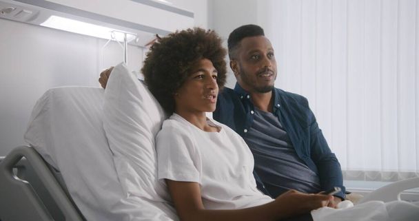Boldog afro-amerikai tinédzser pihen a kórházi szobában, és együtt néz filmet apával. A szobatársak a filmről beszélgetnek. A beteg teljesen felépül a sikeres műtét után. Modern fényes orvosi központ. - Fotó, kép