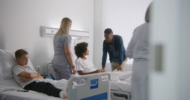 Dojrzały lekarz bandaże nogi młodego chłopca. Afroamerykanin odwiedza syna na oddziale szpitalnym. Pielęgniarka przychodzi do nastolatka i podnosi się z powrotem z łóżka. Pacjent w pełni zdrowy po udanej operacji. - Zdjęcie, obraz