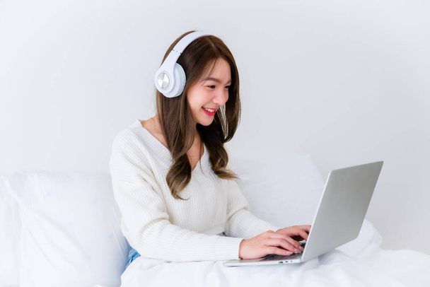 ベッドルームの朝の時間 若いアジアの女性 コンピュータラップトップでハッピー作業, ヘッドセットを着用, 家でベッドに座る - 写真・画像