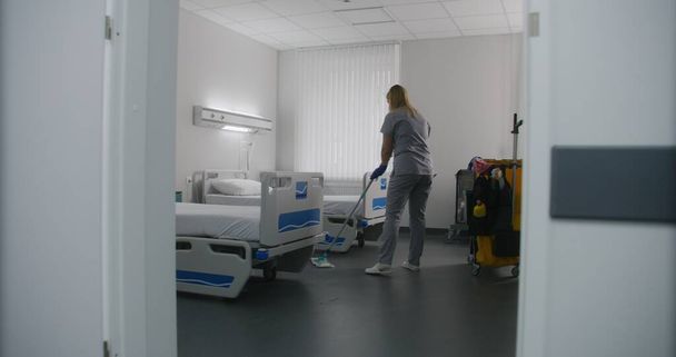 大人の看護師は,病院区のベッドの間の床をモップします. 医療従事者はトロリーを病院の部屋に掃除する. 女性のクリーナーは新しい患者のための病棟を準備します. 近代クリニックで働く医療スタッフ. - 写真・画像