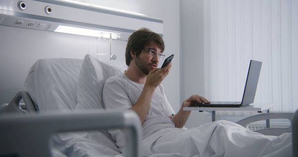 Dorosły mężczyzna leży w łóżku na oddziale szpitalnym, zdalnie pracuje przy użyciu laptopa. Programista pracuje w centrum medycznym i rozmawia przez telefon. Pacjent w pełni zdrowy po udanej operacji. Nowoczesna jasna klinika. - Zdjęcie, obraz