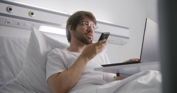 Dorosły mężczyzna leży w łóżku na oddziale szpitalnym i rozmawia z klientem przez telefon. Człowiek w okularach pracuje zdalnie w centrum medycznym. Pacjent w pełni zdrowy po udanej operacji. Nowoczesna jasna klinika. - Zdjęcie, obraz