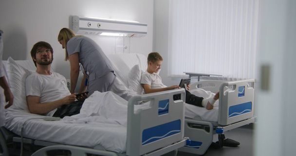 包帯された足を持つ少年はベッドの上に横たわり,タブレットを使用して両親と話します. 女性医師は,ベッドコントローラーの使い方を説明します. 成功した手術後に完全に回復する患者. 現代病院. - 写真・画像