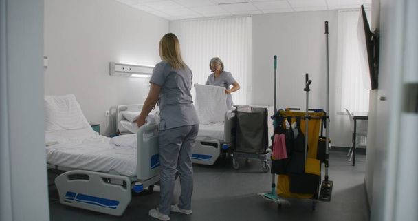 Adult cleaner mops floor with broom in hospital room. Mature health worker changes bedclothes after patients. Nurses clean hospital ward and talk. Medical staff at work in modern medical center. - Photo, Image