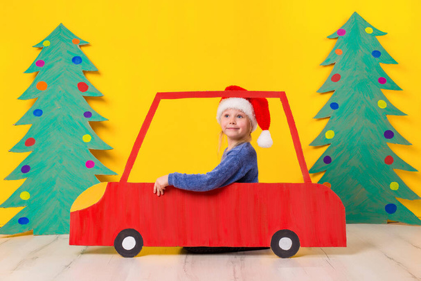 Kind im roten Weihnachtsauto. ein lackiertes Auto und ein grüner Weihnachtsbaum aus Pappe auf gelbem Hintergrund. Weihnachtsferien-Konzept. Kleines emotionales Mädchen - Foto, Bild