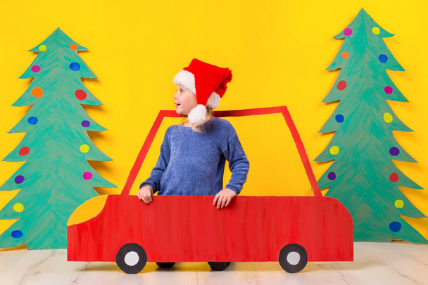 Kind in rode auto van Kerstmis. Een geschilderde auto en de groene kerstboom gemaakt van karton op een gele achtergrond. Xmas concept van de vakantie. Emotionele meisje - Foto, afbeelding