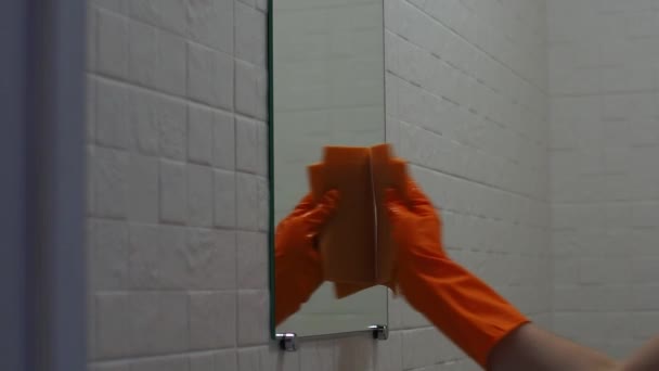 Puhdistus kylpyhuone peili pesuaineella oranssi kumihanskat sienellä. Kodinhoito, siivous, hygienia kodin käsite. Käsi pitää oranssia sientä valkoisella vaahdolla ja pesee peilin. - Materiaali, video