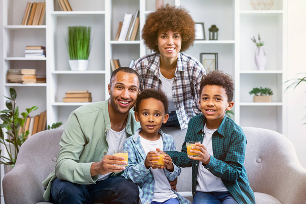 Wesoła afrykańska rodzina spogląda w kamerę. Szczęśliwa matka, ojciec i dwaj synowie piją sok pomarańczowy w salonie i bawią się razem. - Zdjęcie, obraz