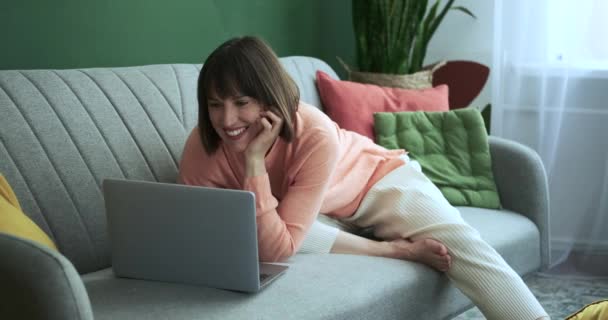 Vuonna viihtyisä olohuone, nainen istuu sohvalla, hänen naurua täyttämällä tilaa, kun hän työskentelee hänen kannettava tietokone. Hänen tarttuva ilonsa tuo iloa ja positiivisuutta huoneeseen.. - Materiaali, video