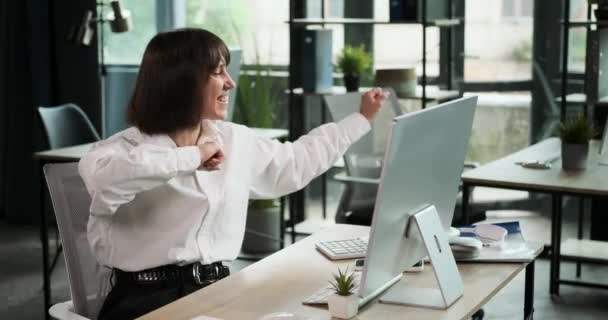 Eine fröhliche kaukasische Unternehmerin kann ihr Glück nicht fassen und beginnt zu tanzen, während sie an ihrem Schreibtisch im Büro sitzt. Ihr lebhafter Tanz spiegelt ihre Begeisterung wider. - Filmmaterial, Video